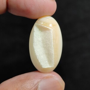 May include: A white and beige agate cabochon with a natural crystal formation inside. The cabochon is oval shaped and has a smooth, polished surface.