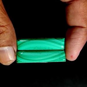 May include: Two rectangular malachite gemstones, held between fingers against a black background. The stones display a vibrant green color with swirling patterns. The polished surfaces reflect light, showcasing the natural beauty of the mineral.
