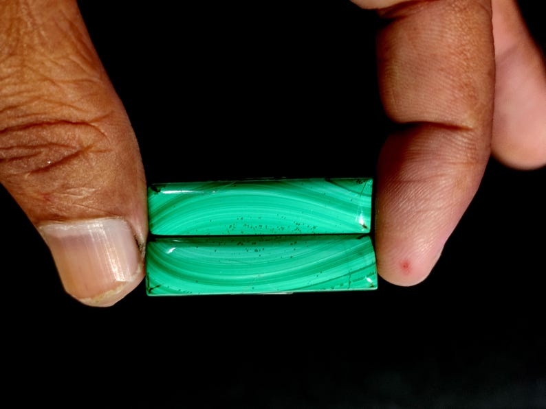 May include: Two rectangular malachite gemstones, held between fingers against a black background. The stones display a vibrant green color with swirling patterns. The polished surfaces reflect light, showcasing the natural beauty of the mineral.