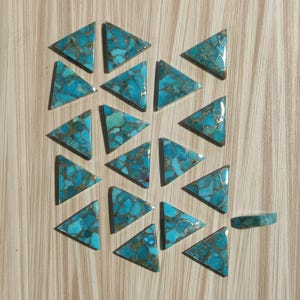 Puede incluir: Una colección de 19 cabujones de turquesa en forma de triángulo con motas doradas. Los cabujones están dispuestos en un patrón circular sobre una superficie de madera.