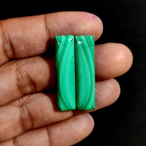 May include: Two rectangular malachite stones with a vibrant green color and distinctive swirling patterns. The stones are displayed against a dark background, highlighting their natural beauty and unique veining.