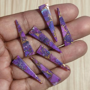 May include: A collection of eight purple and gold triangular shaped gemstone cabochons. The cabochons are approximately 1 inch in length and have a smooth, polished surface.