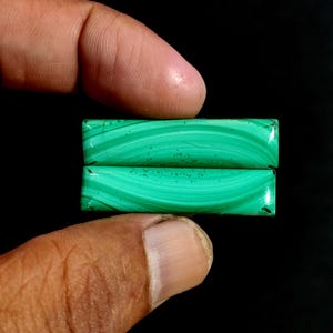 May include: Two rectangular malachite gemstones, each displaying a vibrant green color with distinctive swirling patterns. The stones are held against a black background, highlighting their natural beauty and unique veining.