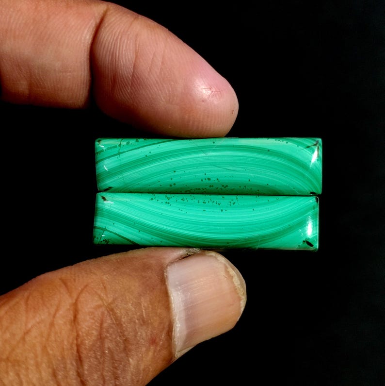 May include: Two rectangular malachite gemstones, each displaying a vibrant green color with distinctive swirling patterns. The stones are held against a black background, highlighting their natural beauty and unique veining.