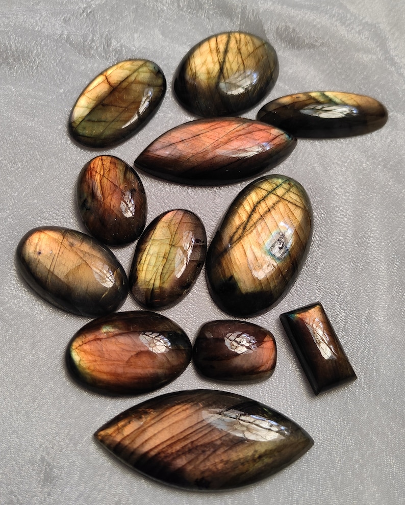 May include: A collection of 11 labradorite gemstone cabochons in various shapes and sizes. The stones are a mix of dark green, black, and gold colors with iridescent flashes.