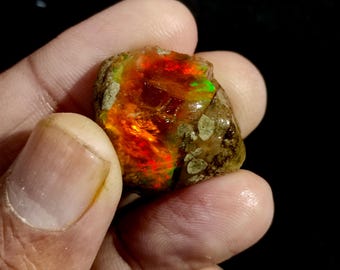 May include: A rough, natural opal gemstone with a mix of orange, red, and green colors. The opal is held in a hand.