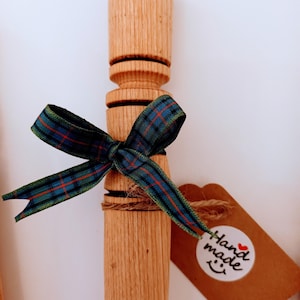 May include: A wooden rolling pin with a blue and green tartan ribbon tied around it. The rolling pin has a hand-carved design and is tied with twine. A brown tag with the text "hand made" is attached to the ribbon.