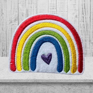 Könnte beinhalten: Ein Filz-Patch in Form eines Regenbogens mit einem lila Herz in der Mitte. Der Regenbogen hat rote, orange, gelbe, grüne, blaue und lila Streifen.