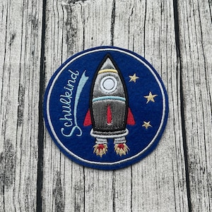Könnte beinhalten: Ein blauer Filz-Kreis mit einer silbernen gestickten Rakete und dem Wort "Schulkind" in Weiß. Die Rakete hat rote Flammen und ein weißes Fenster. Im Hintergrund befinden sich drei gelbe Sterne.