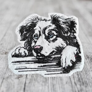 Könnte beinhalten: Gestickter Aufnäher mit einem schwarz-weißen Hund mit blauen Augen und einer rosa Nase. Der Kopf und die Pfoten des Hundes sind sichtbar, als würde er über eine Oberfläche schauen. Der Aufnäher hat einen weißen Rand und ist vor einem Holzgrund platziert.