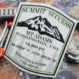 May include: A rectangular sticker featuring a black mountain graphic, the text SUMMIT SUCCESS, MT ADAMS, WASHINGTON, USA, 12,280 FT, 46.2024 N, -121.4910 W, and FORTUNE FAVORS THE BOLD, framed by a green border, positioned between orange and silver carabiners.