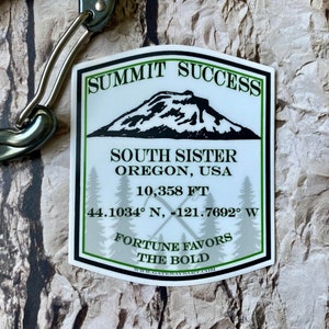 May include: White sticker with green text and a mountain silhouette. The text reads "Summit Success", "South Sister", "Oregon, USA", "10,358 FT", "44.1034° N, -121.7692° W", "Fortune Favors The Bold", and "www.oregonwaysigns.com".