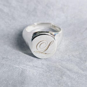 Peut inclure: Une bague chevalière en argent avec une finition polie. La bague présente une face circulaire gravée de la lettre "L" dans une écriture élégante. Un bijou classique.