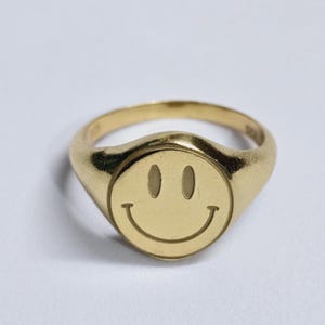 May include: A gold ring featuring a smiley face design. The ring has a smooth, polished finish and a rounded band. The smiley face is centered on a circular face. The ring is a simple, yet stylish piece of jewellery.
