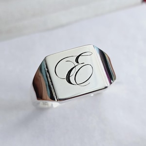 Peut inclure: Une bague chevalière en argent avec une face carrée. La bague présente une lettre "E" en cursive gravée sur la face. La bague est présentée dans un écrin rouge et blanc, suggérant qu'il s'agit d'un bijou.