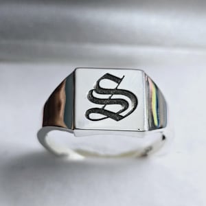 Peut inclure: Une bague chevalière en argent avec une face carrée. La face présente un "S" noir stylisé dans une police ornée. La bague a une finition polie et un design classique.