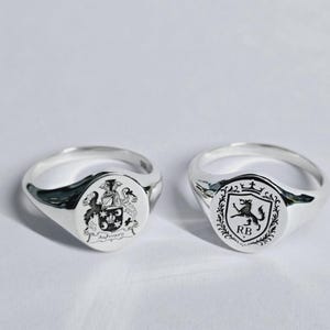 Peut inclure: Deux chevalières en argent avec des armoiries gravées. L'une présente un blason familial détaillé avec le nom "Aylward". L'autre affiche un blason avec un lion et les initiales "RB".