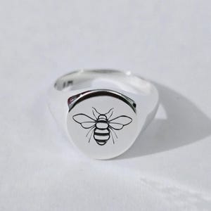 Peut inclure: Bague chevalière argentée avec un motif d'abeille noire détaillé sur la face ovale. La bague est en métal poli et brillant, posée sur une surface blanche. Le dessin de l'abeille comprend des ailes et un abdomen rayé. La bague mesure environ 1,8 cm.