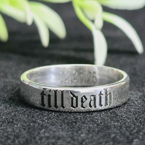 Könnte beinhalten: Ein silberner Ring mit dem eingravierten Schriftzug "till death" in gotischer Schrift. Der Ring trägt auch das Datum "12.25.25". Der Ring liegt auf einer schwarzen Oberfläche mit einem verschwommenen grün-weißen Hintergrund.