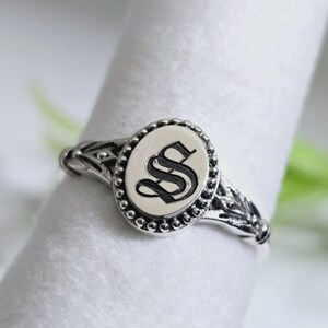 May include: Silver ring with an oval face displaying the letter "S" in black script. The ring has a beaded border and decorative detailing on the band. The ring is shown on a white surface.