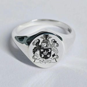 May include: A polished silver signet ring, featuring a detailed family crest. The crest includes a shield with intricate designs, a banner reading "Aylward", and decorative elements. The ring has a classic, rounded band.