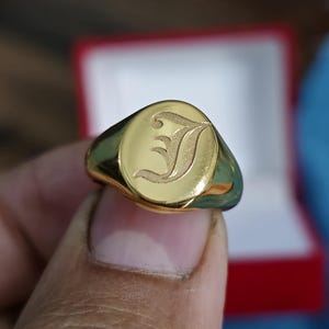 May include: A gold signet ring with a polished finish. The ring features an engraved letter "J" on an oval face. The ring is held in a hand, with a red and white box in the background.