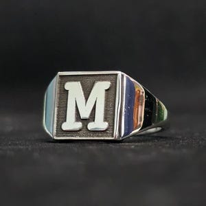 May include: A silver-toned signet ring featuring a rectangular face with a polished, raised letter M centered against a dark, textured background. The band of the ring is smooth and reflective, showcasing a metallic finish with clean, sharp edges.
