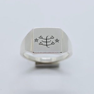 Peut inclure: Une bague chevalière en argent avec une face carrée. La face présente un motif graphique noir avec une forme stylisée centrale et quatre étoiles environnantes. La bague est présentée sur fond blanc.