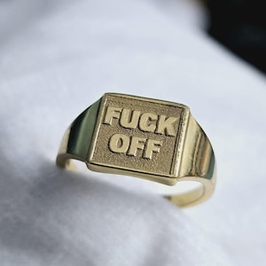 Peut inclure: Une bague chevalière dorée avec les mots "FUCK OFF" en relief sur une face carrée. La bague a une finition polie et un anneau simple.