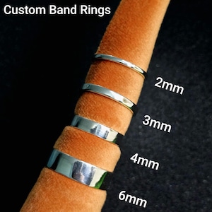Peut inclure: Quatre bagues en métal argenté présentées sur un support conique en velours orange. Les bagues sont étiquetées avec des largeurs de 2mm, 3mm, 4mm et 6mm. Le texte Custom Band Rings est visible en haut à gauche, indiquant le type de produit.