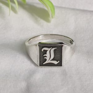 May include: A silver signet ring with a square face featuring the letter "L" in a gothic font. The ring is made of polished metal and sits on a white surface. The ring is a classic piece of jewelry.