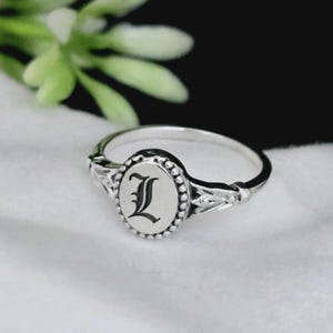 May include: A silver ring with an oval face displaying the letter "L" in a black, stylized font. The ring has a beaded border and decorative flourishes on the band.