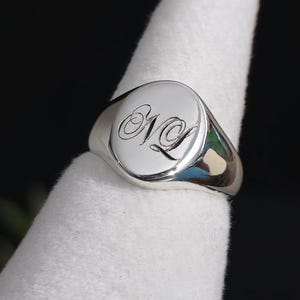Può includere: Un anello con sigillo in argento lucido con una superficie ovale liscia. La superficie dell'anello presenta le iniziali "NL" incise in una scrittura elegante. L'anello è mostrato su una superficie bianca, con uno sfondo scuro.