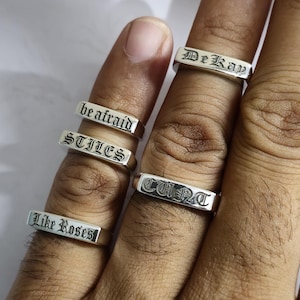 May include: Five silver-toned metal rings featuring engraved text in a black gothic-style font. The rings display the words DeKay, be afraid, STILES, CUNT, and Like Roses. Each ring has a flat, rectangular face with the lettering centred.