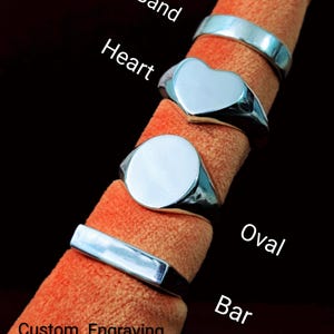 May include: Four silver-toned rings with smooth, polished surfaces. The rings are labelled as Band, Heart, Oval, and Bar. The text Custom Engraving Available is printed at the bottom. Each ring features a distinct shape, including a simple band, a heart-shaped face, a circular face, and a rectangular bar face.