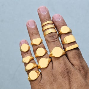 May include: A collection of gold-colored signet rings in various shapes, including round, oval, heart, and rectangular designs. The rings are displayed on fingers against a light blue background.