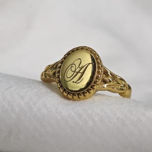 Peut inclure: Une bague chevalière dorée avec une face ovale. La face présente la lettre "A" stylisée en écriture cursive. La bague a une bordure perlée autour de la face et des détails décoratifs sur la bande.