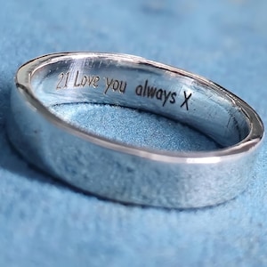 May include: A silver ring with the inscription "21 Love you always X" engraved inside. The ring has a smooth, polished finish and a rounded design. The background is a soft blue. The ring's width is approximately 0.3 cm.