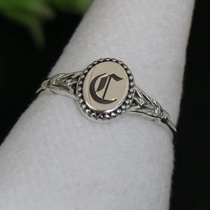 Puede incluir: Un anillo de plata con una cara ovalada que presenta la letra "C" en una fuente serif. El anillo tiene un diseño detallado en la banda y alrededor de la cara, con un borde de cuentas. El anillo se muestra sobre una superficie blanca.