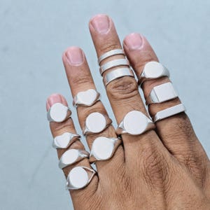 Peut inclure: Plusieurs bagues chevalières en argent de formes variées, dont ovales, carrées et en forme de cœur. Les bagues sont présentées sur des doigts sur un fond bleu clair. Les bagues sont en métal brillant et réfléchissant.