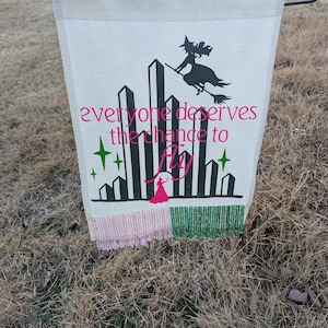 Pode incluir: Uma bandeira de jardim branca com uma ilustração verde, preta e branca da Cidade Esmeralda do Mágico de Oz. O texto "everyone deserves the chance to fly" está impresso em rosa na bandeira. A bandeira tem franjas rosa e verdes.