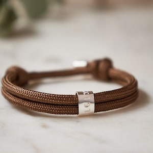 Peut inclure: Un bracelet en cordon double brin marron avec un charme personnalisé en argent qui dit "Lex".