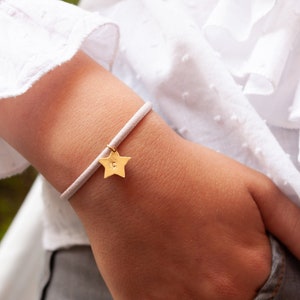 Könnte beinhalten: Ein weißes elastisches Armband mit einem goldenen Sternanhänger. Der Stern hat einen kleinen gravierten Buchstaben "C".