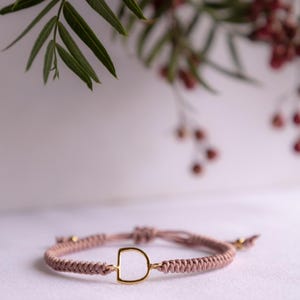 May include: A mauve braided cord bracelet featuring a central gold-toned metal letter D charm. The bracelet includes adjustable sliding knots at the back, finished with small gold-toned beads at the ends of the cord, creating a simple, functional design.