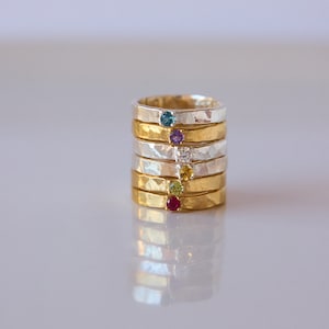 Pode incluir: Uma pilha de seis anéis de ouro e prata com pedras preciosas de diferentes cores. Os anéis estão empilhados uns sobre os outros, com o anel inferior de ouro e o anel superior de prata. As pedras preciosas são vermelha, verde, amarela, branca, roxa e azul.