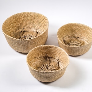 May include: Three woven seagrass baskets of varying sizes, all with a natural beige colour. The baskets are round and have a slight lip around the top.
