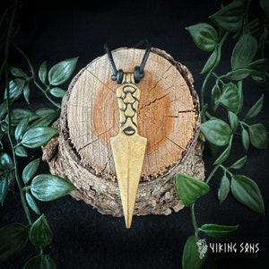May include: A gold-coloured pendant shaped like a spearhead with intricate details. The pendant is hanging from a black leather cord and is resting on a piece of wood.