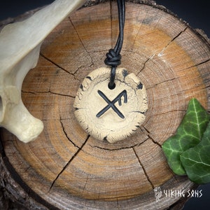 May include: A white ceramic pendant with a black Viking rune symbol on a black leather cord. The pendant is on a wooden surface.