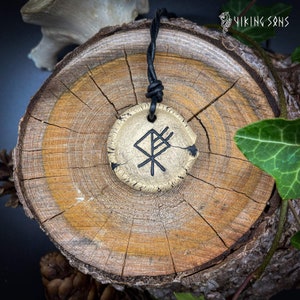 May include: A black leather cord necklace with a round, cream-coloured pendant featuring a black Vegvisir symbol. The pendant is on a wooden surface.