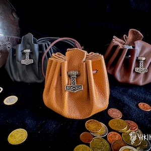 Puede incluir: Tres bolsas de cuero en negro, marrón y beige, cada una adornada con un emblema plateado del martillo de Thor. Las bolsas se cierran en la parte superior con cordones. Varias monedas están esparcidas.